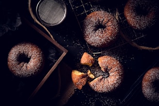 A variety of powdered sugar-covered donuts are displayed on a black surface. Some are placed on a cooling rack, while others are inside a wooden box. A broken donut reveals its soft, golden interior. A metal sifter is nearby, suggesting the process of sprinkling the sugar.