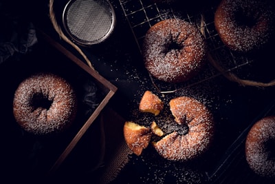 A variety of powdered sugar-covered donuts are displayed on a black surface. Some are placed on a cooling rack, while others are inside a wooden box. A broken donut reveals its soft, golden interior. A metal sifter is nearby, suggesting the process of sprinkling the sugar.
