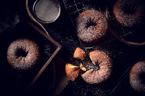 A variety of powdered sugar-covered donuts are displayed on a black surface. Some are placed on a cooling rack, while others are inside a wooden box. A broken donut reveals its soft, golden interior. A metal sifter is nearby, suggesting the process of sprinkling the sugar.