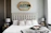 A well-designed bedroom featuring a large upholstered bed with a tufted headboard. The bed is adorned with textured and patterned pillows. On either side of the bed are wooden nightstands, each holding a silver lamp. Above the bed is an ornate mirror with a gold frame. Decorative geometric accents and a soft gray throw blanket complete the room's elegant and cozy atmosphere.