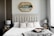 A well-designed bedroom featuring a large upholstered bed with a tufted headboard. The bed is adorned with textured and patterned pillows. On either side of the bed are wooden nightstands, each holding a silver lamp. Above the bed is an ornate mirror with a gold frame. Decorative geometric accents and a soft gray throw blanket complete the room's elegant and cozy atmosphere.