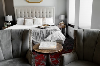A stylish and cozy bedroom features a large, cushioned bed with decorative pillows and a textured throw blanket. The room includes two elegant armchairs with a wooden round table positioned between them. On the table is a tray with a glass bottle and glasses. The room is decorated with soft lighting from two bedside lamps, and the overall color scheme is neutral with hints of gray and green.