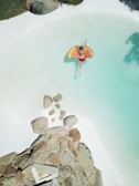 A person in a red swimsuit floats on an orange slice-shaped inflatable in a light blue swimming pool. Large rocks are positioned at the edge of the pool, and there is surrounding greenery.