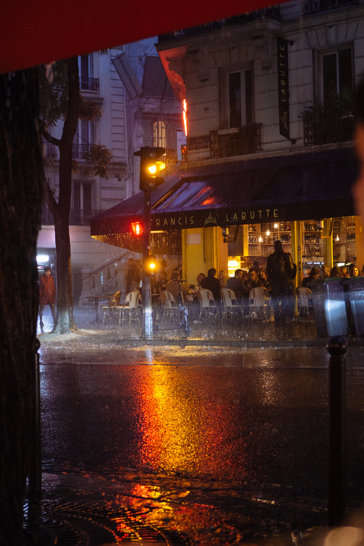 A lit café terrace on a rainy night, warm light reflected across the wet street.