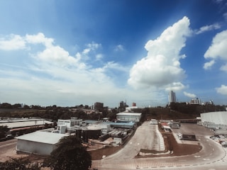A wide-angle view of an industrial area with several large factory buildings and roads surrounded by greenery. In the distance, a city skyline with high-rise buildings beneath a partly cloudy blue sky. The scene combines both urban and natural elements.