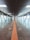 A long, enclosed pedestrian walkway with large ceiling lights and glass windows on both sides. Several people are walking along the path, which has a central tactile guide strip on the floor.
