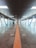 A long, enclosed pedestrian walkway with large ceiling lights and glass windows on both sides. Several people are walking along the path, which has a central tactile guide strip on the floor.