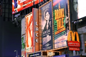 Billboards and advertisements in a vibrant urban setting showcasing theater show posters and illuminated signs. Among them, there are posters for 'A Bronx Tale' and 'Sunset Boulevard,' with bright lights and a commercial sign for a fast-food chain visible.
