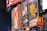 Billboards and advertisements in a vibrant urban setting showcasing theater show posters and illuminated signs. Among them, there are posters for 'A Bronx Tale' and 'Sunset Boulevard,' with bright lights and a commercial sign for a fast-food chain visible.