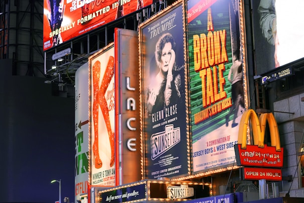 Billboards and advertisements in a vibrant urban setting showcasing theater show posters and illuminated signs. Among them, there are posters for 'A Bronx Tale' and 'Sunset Boulevard,' with bright lights and a commercial sign for a fast-food chain visible.