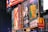 Billboards and advertisements in a vibrant urban setting showcasing theater show posters and illuminated signs. Among them, there are posters for 'A Bronx Tale' and 'Sunset Boulevard,' with bright lights and a commercial sign for a fast-food chain visible.