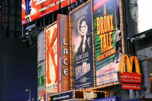 Billboards and advertisements in a vibrant urban setting showcasing theater show posters and illuminated signs. Among them, there are posters for 'A Bronx Tale' and 'Sunset Boulevard,' with bright lights and a commercial sign for a fast-food chain visible.