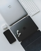 A collection of sleek electronic devices, including a laptop, tablet, smartphone, earbuds, keyboard, and charging case, arranged on a white surface. The devices are predominantly silver and black, conveying a modern and minimalist aesthetic.