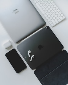 A collection of sleek electronic devices, including a laptop, tablet, smartphone, earbuds, keyboard, and charging case, arranged on a white surface. The devices are predominantly silver and black, conveying a modern and minimalist aesthetic.