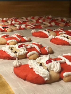 A collection of Santa Claus-themed cookies decorated with red and white icing. The cookies are shaped and decorated to resemble Santa's face, complete with a white beard and red hat. Some have chocolate chip eyes and a red candy nose, resembling a festive scene laid out on a baking sheet.