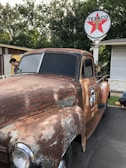 A rusty vintage pickup truck is parked in front of a Texaco gas station. The truck shows signs of wear and aging, with a weathered paint job and oxidized surfaces. Above, a Texaco sign with a red star logo is mounted on a pole. Surrounding the scene are trees with lush foliage, and a person is seen next to the truck examining it.