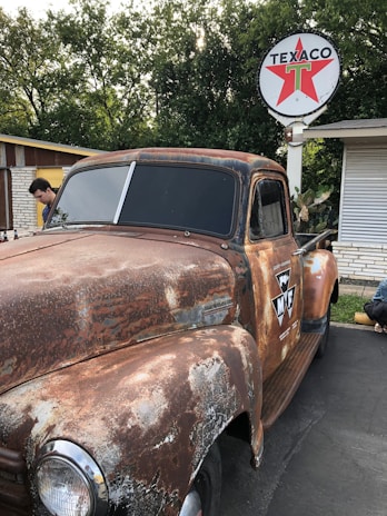 A rusty vintage pickup truck is parked in front of a Texaco gas station. The truck shows signs of wear and aging, with a weathered paint job and oxidized surfaces. Above, a Texaco sign with a red star logo is mounted on a pole. Surrounding the scene are trees with lush foliage, and a person is seen next to the truck examining it.
