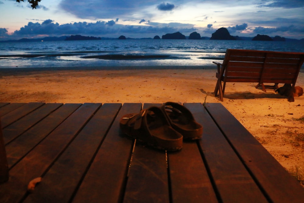 sandálias sobre mesa de madeira perto do banco de frente para o oceano durante a hora dourada