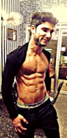 A fit man with visible abdominal muscles and a slight smile poses in an indoor setting, wearing dark pants and an open jacket. The background features a wall with patterned wallpaper and glass doors.