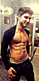 A fit man with visible abdominal muscles and a slight smile poses in an indoor setting, wearing dark pants and an open jacket. The background features a wall with patterned wallpaper and glass doors.