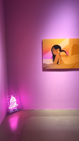 A minimalistic art gallery room with pink walls features a framed artwork of a stylized figure in warm colors, predominantly oranges and pinks. Near the corner, a neon light sculpture resembling an ice cream cone glows in bright pink.