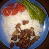 A plate of white rice served with cooked meat pieces, garnished with slices of fresh tomatoes and onions, accompanied by green leafy vegetables on the side. The meal is presented on a blue plate.