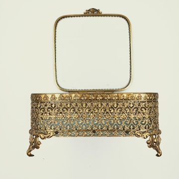 An ornate, antique-style jewelry box with intricate gold filigree design and a decorative border. The box is elevated on elegant, claw-like feet and the lid is open, revealing a square mirror with a delicate frame.