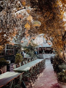 An outdoor dining area featuring long wooden tables with chairs, surrounded by lush foliage. Ornate glass lanterns hang from branches overhead, casting a warm glow. The setting has a rustic bar made of red brick with a sign that reads 'The Garden Bar'. The atmosphere is cozy and inviting, reminiscent of a secluded garden retreat.