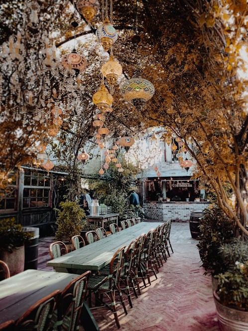 An outdoor dining area featuring long wooden tables with chairs, surrounded by lush foliage. Ornate glass lanterns hang from branches overhead, casting a warm glow. The setting has a rustic bar made of red brick with a sign that reads 'The Garden Bar'. The atmosphere is cozy and inviting, reminiscent of a secluded garden retreat.