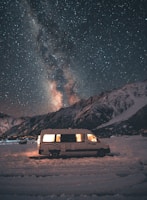 A camper van is parked on a snowy landscape with a backdrop of a dark, star-filled sky. The Milky Way is visible, creating a breathtaking scene. The van's interior is softly lit, adding warmth against the cold, vast night sky and rugged mountains.