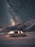 A camper van is parked on a snowy landscape with a backdrop of a dark, star-filled sky. The Milky Way is visible, creating a breathtaking scene. The van's interior is softly lit, adding warmth against the cold, vast night sky and rugged mountains.