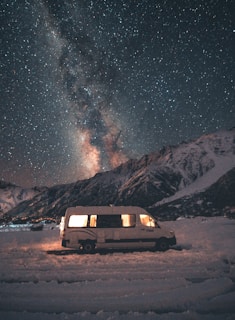 A camper van is parked on a snowy landscape with a backdrop of a dark, star-filled sky. The Milky Way is visible, creating a breathtaking scene. The van's interior is softly lit, adding warmth against the cold, vast night sky and rugged mountains.