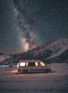 A camper van is parked on a snowy landscape with a backdrop of a dark, star-filled sky. The Milky Way is visible, creating a breathtaking scene. The van's interior is softly lit, adding warmth against the cold, vast night sky and rugged mountains.