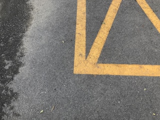 A section of asphalt pavement marked with bright yellow painted lines, creating a geometric pattern. Some small bits of debris and leaves are scattered on the surface. The pavement appears slightly textured and weathered.