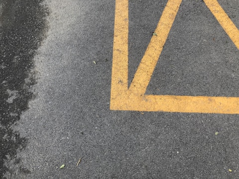 A section of asphalt pavement marked with bright yellow painted lines, creating a geometric pattern. Some small bits of debris and leaves are scattered on the surface. The pavement appears slightly textured and weathered.