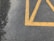 A section of asphalt pavement marked with bright yellow painted lines, creating a geometric pattern. Some small bits of debris and leaves are scattered on the surface. The pavement appears slightly textured and weathered.