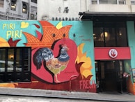 A colorful mural featuring a large, detailed rooster with a vibrant feather pattern. Bright colors such as red, orange, teal, and yellow dominate the mural, which also includes text like 'Piri Piri', 'our passion', and 'fresh'. The mural is on the side of a building, next to the entrance of a restaurant with a red logo displaying a rooster.