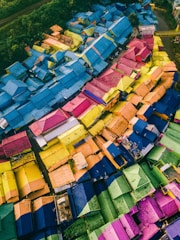 Aerial view of a densely packed urban neighborhood featuring an array of vividly painted rooftops in a mosaic pattern. The roofs are painted in bright colors including blue, yellow, pink, green, and orange, creating a visually striking patchwork effect. The scene is surrounded by lush greenery, indicating that the area is adjacent to natural vegetation.