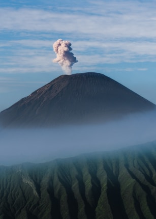 A volcanic mountain with a plume of smoke rising from its summit, surrounded by a lush green landscape and a layer of mist. The sky above is clear with some clouds.