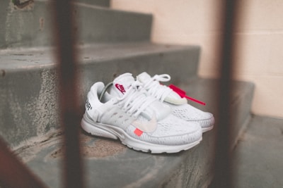 A pair of white athletic shoes with red accents is placed on a set of concrete steps. The shoes have distinct branding and are laced, with one shoe featuring a visible tag.
