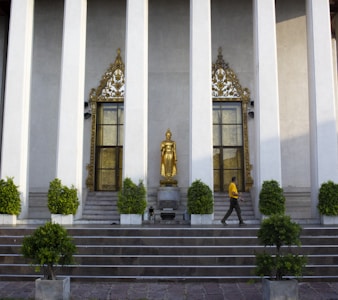 A serene temple facade featuring tall columns and intricate golden decorations framing two windows. In front of the building stands a golden Buddha statue on a pedestal, surrounded by neatly trimmed green shrubs. A person wearing a yellow shirt is walking in front of the temple.