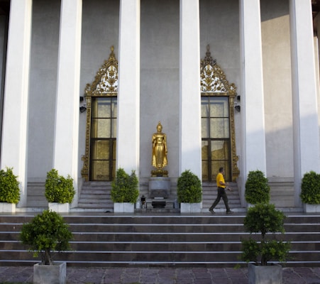A serene temple facade featuring tall columns and intricate golden decorations framing two windows. In front of the building stands a golden Buddha statue on a pedestal, surrounded by neatly trimmed green shrubs. A person wearing a yellow shirt is walking in front of the temple.