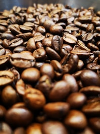 A close-up view of a pile of roasted coffee beans, showcasing their rich brown tones and textured surfaces. The focus is primarily on the beans in the foreground, with more beans fading into the background, creating a sense of depth.