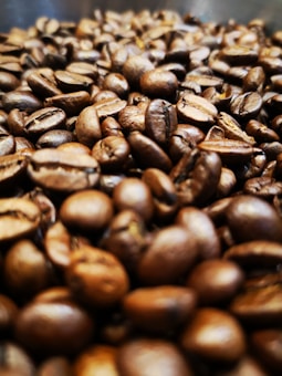 A close-up view of a pile of roasted coffee beans, showcasing their rich brown tones and textured surfaces. The focus is primarily on the beans in the foreground, with more beans fading into the background, creating a sense of depth.