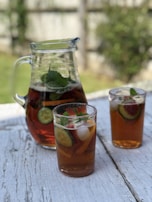 A pitcher filled with iced tea and slices of cucumber, strawberries, and mint leaves is placed on a weathered wooden table. Two glasses, also containing the same beverage with ice cubes and garnishes, are nearby. The background features a blurred garden setting with greenery.
