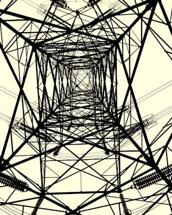 A symmetrical view of a transmission tower from directly beneath, showcasing intricate crisscrossing metal beams and cables. The structure creates complex geometric patterns against a light background.