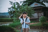 A young boy wearing a school uniform and backpack stands in a rural setting. Behind him is another child whose face is partially obscured. The background features a small wooden house surrounded by lush greenery and a rice paddy field.