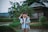 A young boy wearing a school uniform and backpack stands in a rural setting. Behind him is another child whose face is partially obscured. The background features a small wooden house surrounded by lush greenery and a rice paddy field.