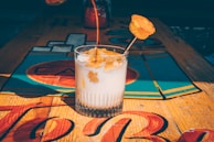 A glass filled with a creamy, white beverage, topped with a caramel-colored syrup being poured. There's a slice of fruit or garnish skewered on a stick resting inside the drink. The glass sits on a colorful, wooden table with bright, abstract designs.