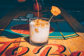A glass filled with a creamy, white beverage, topped with a caramel-colored syrup being poured. There's a slice of fruit or garnish skewered on a stick resting inside the drink. The glass sits on a colorful, wooden table with bright, abstract designs.